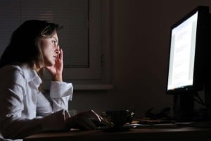 A woman is sitting in the dark squinting at her bright white computer screen.