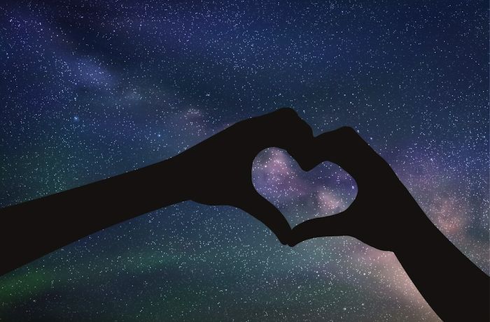 Two hands reflected in black making a heart against a starry night sky.