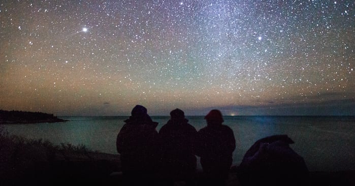 Three people sit looking over the water at Acadia National Park. The night sky is filled with star and you can see a hint of he Milky Way.