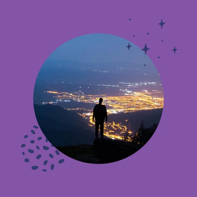 Photo illustration of a person on a hill at night overlooking bright city lights.