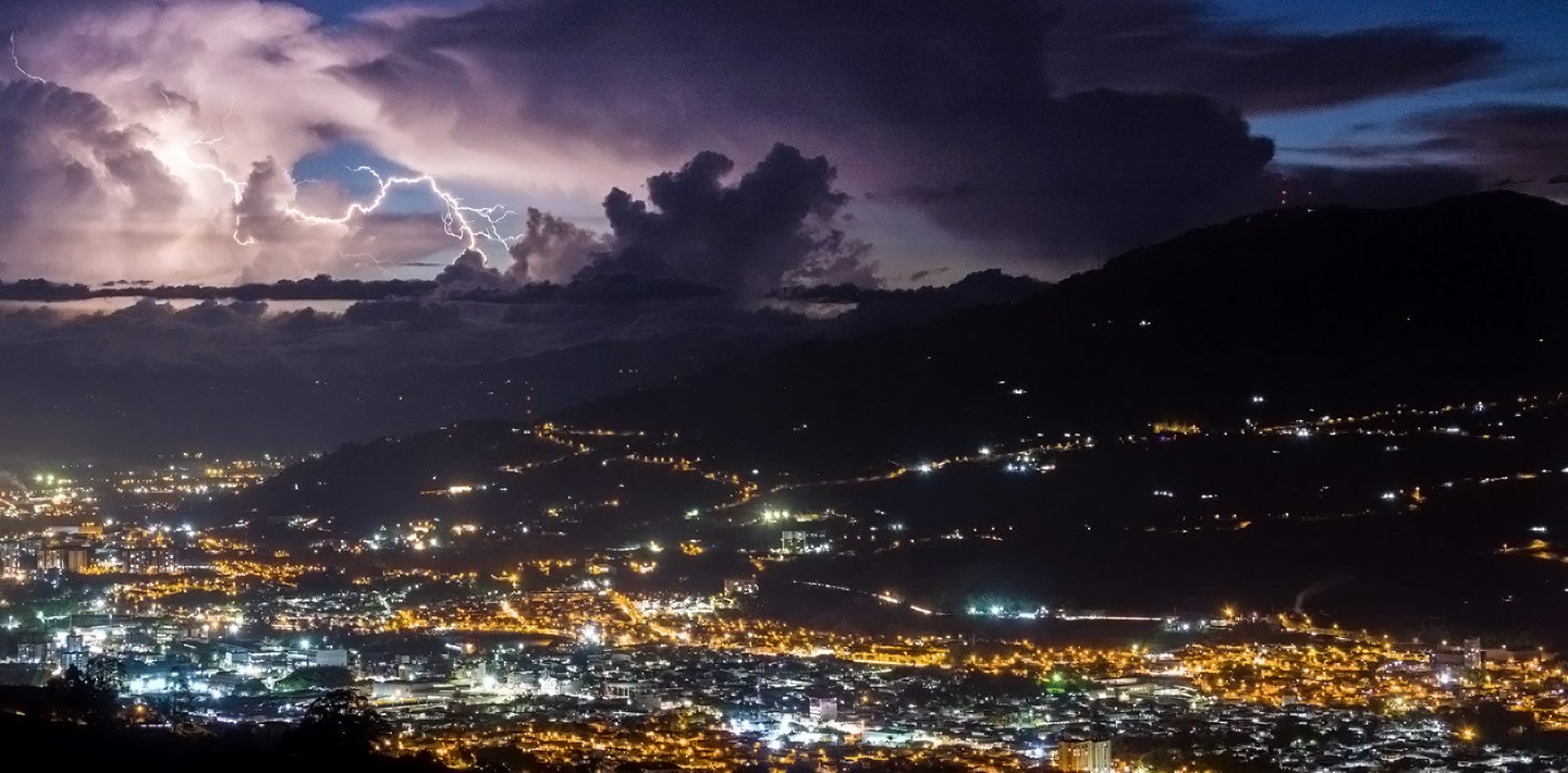 Nighttime photo looking down into a city full of lights, with storm clouds and lightning in the distance.