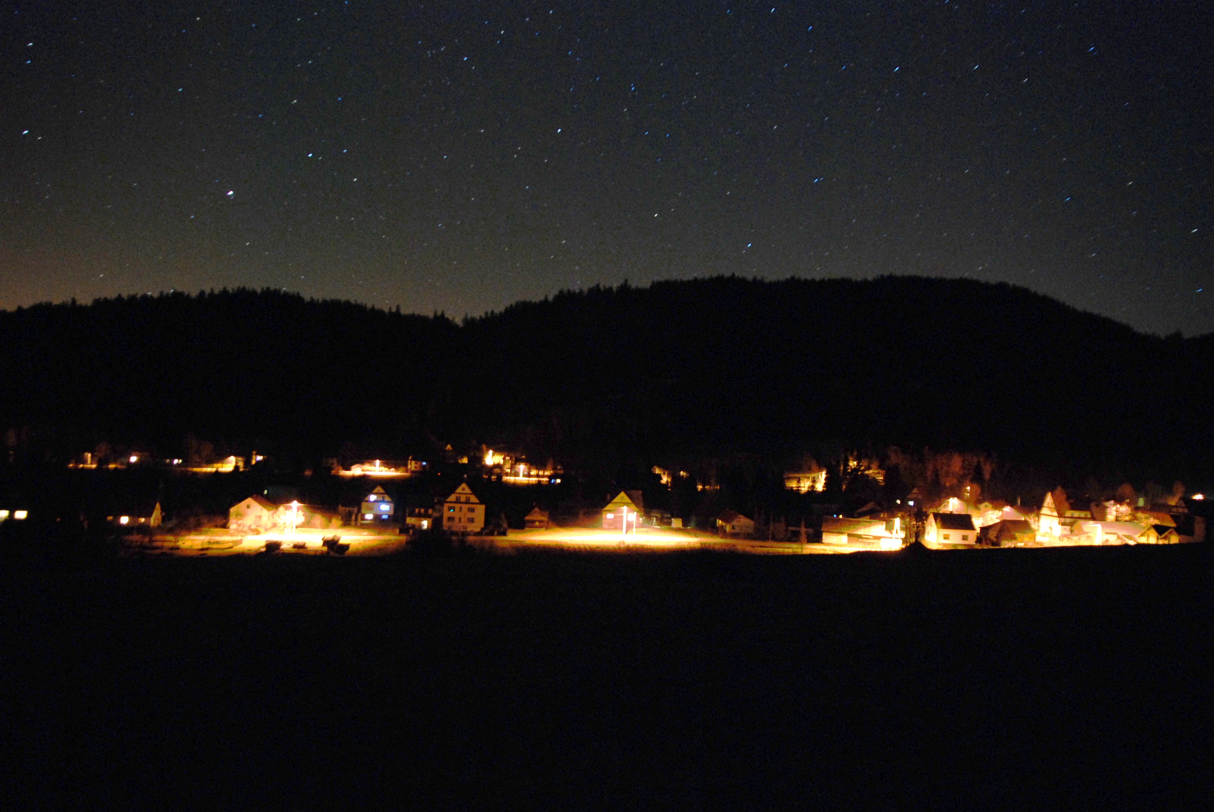 A small neighborhood of houses with warm down-shielded lights illuminating the street, sit below a sky filled with stars.