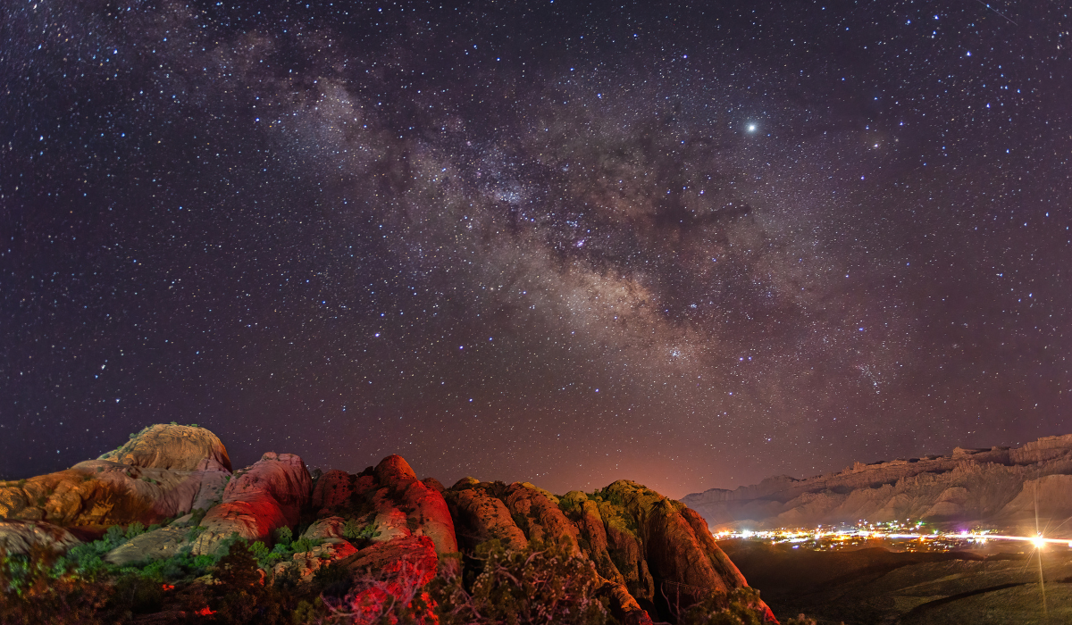 Red rock cliffs give way to city lights with the Milky Way overhead.