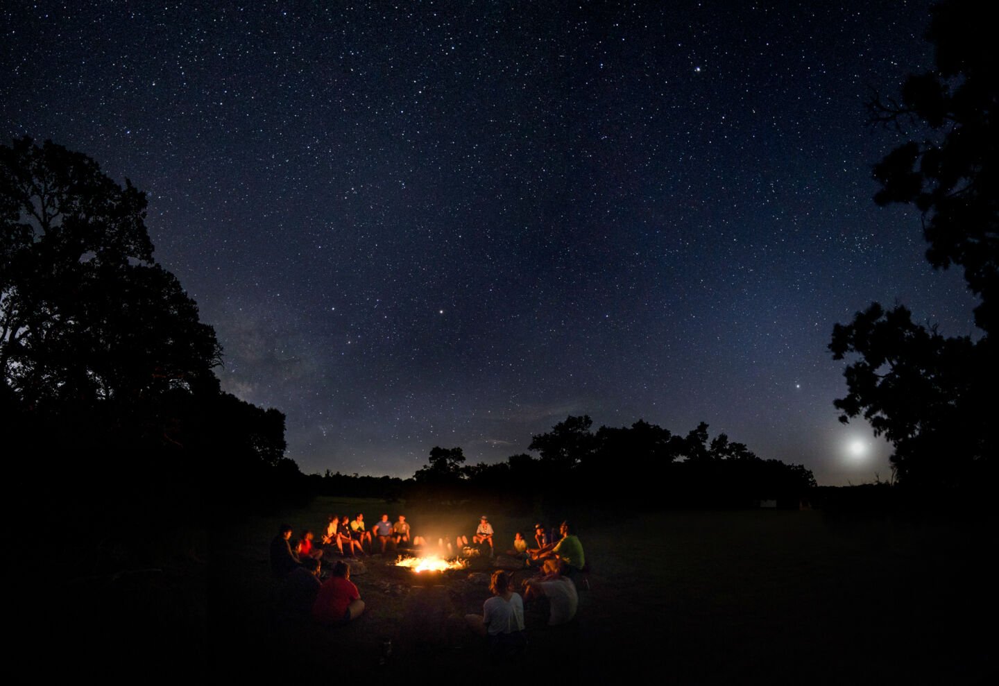 A group of people sit around a campfire under a starry Texas sky.