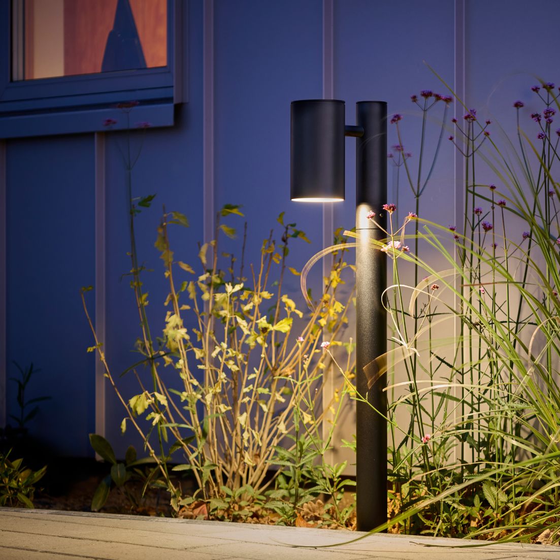 Modern black bollard-style outdoor light illuminating garden plants beside a house at night, casting warm, downward-directed light that highlights surrounding grasses and flowers near a window.