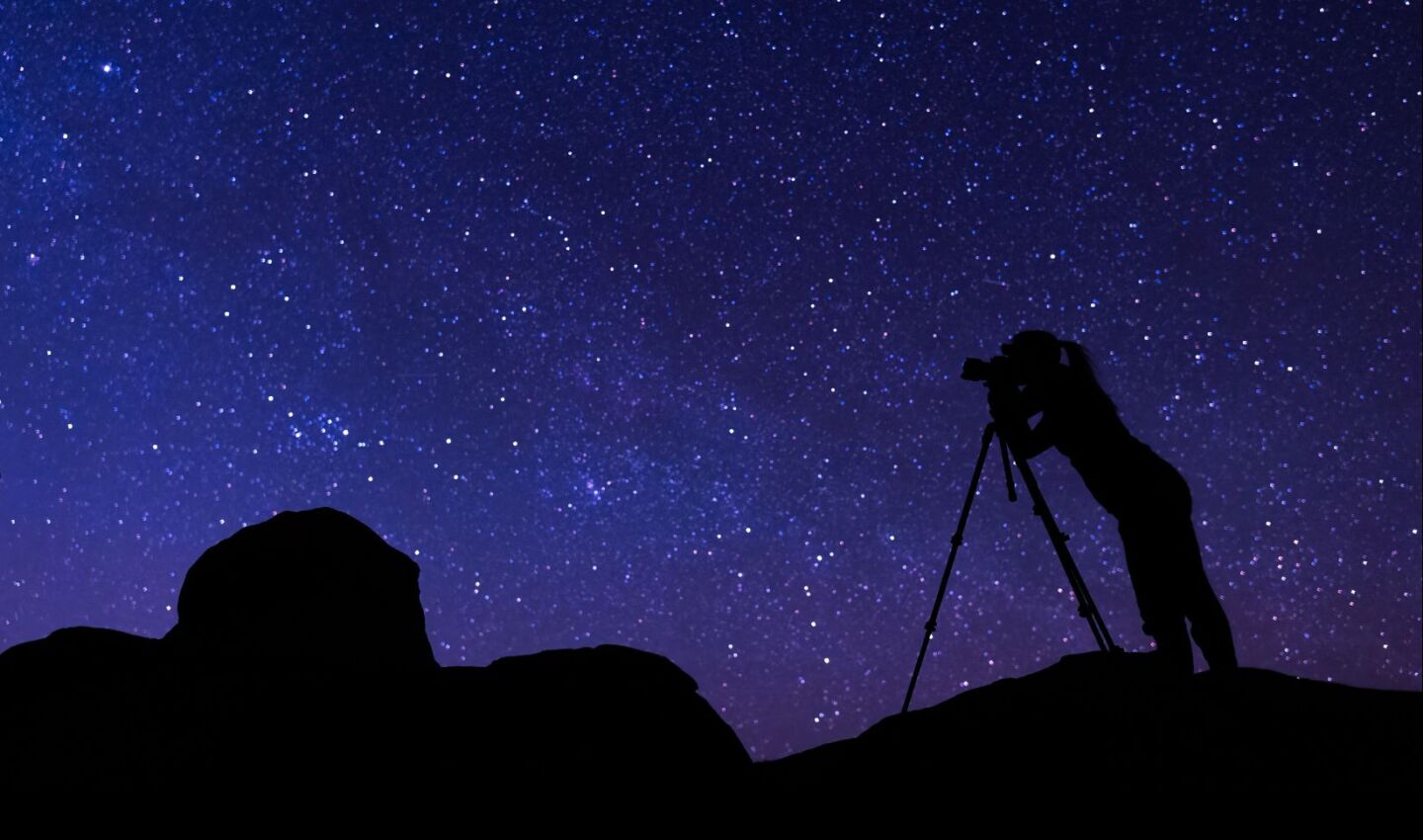 Silhouette of a photographer standing on rocky terrain and looking through a camera on a tripod beneath a dense, star-filled night sky with the Milky Way visible.