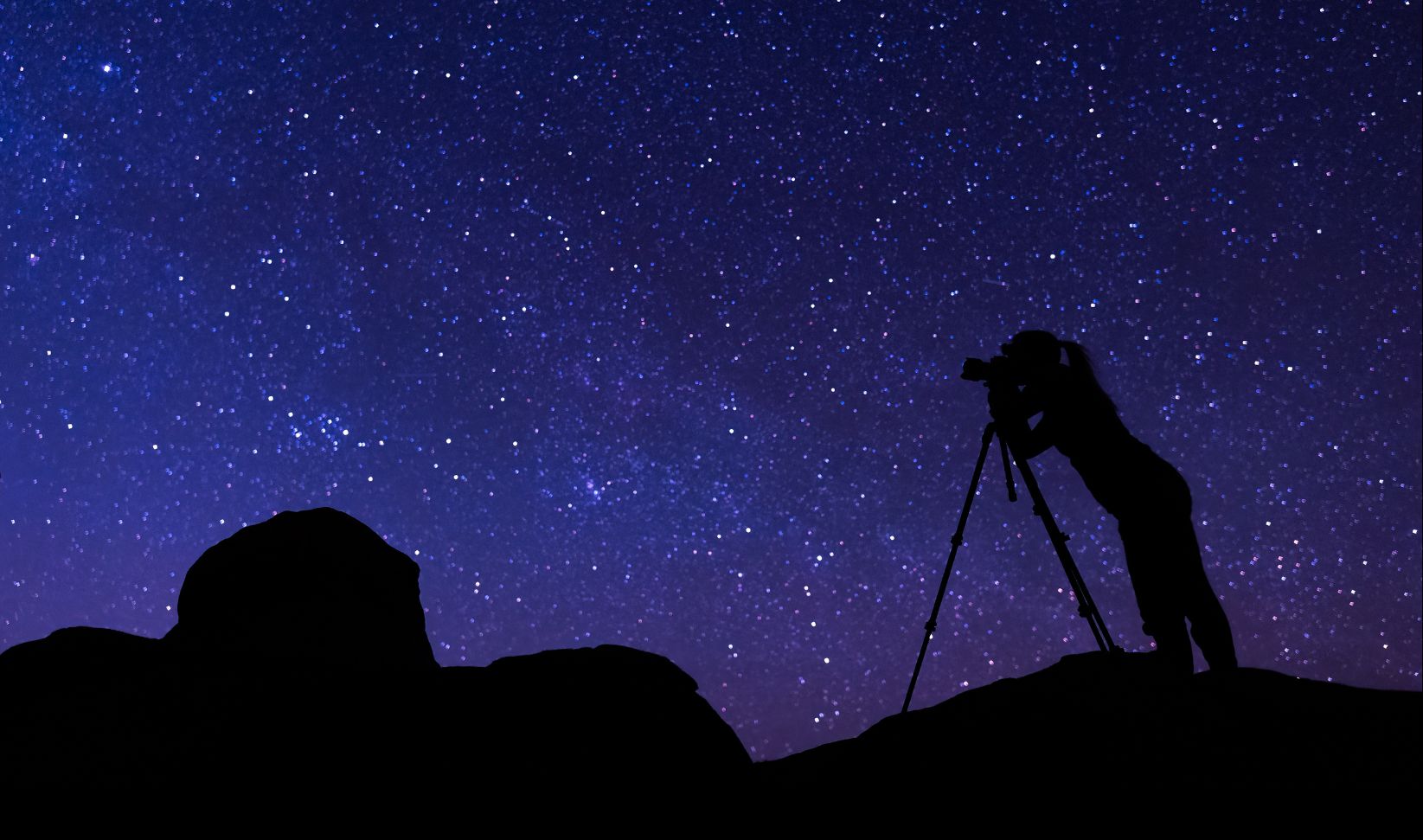 Silhouette of a photographer standing on rocky terrain and looking through a camera on a tripod beneath a dense, star-filled night sky with the Milky Way visible.
