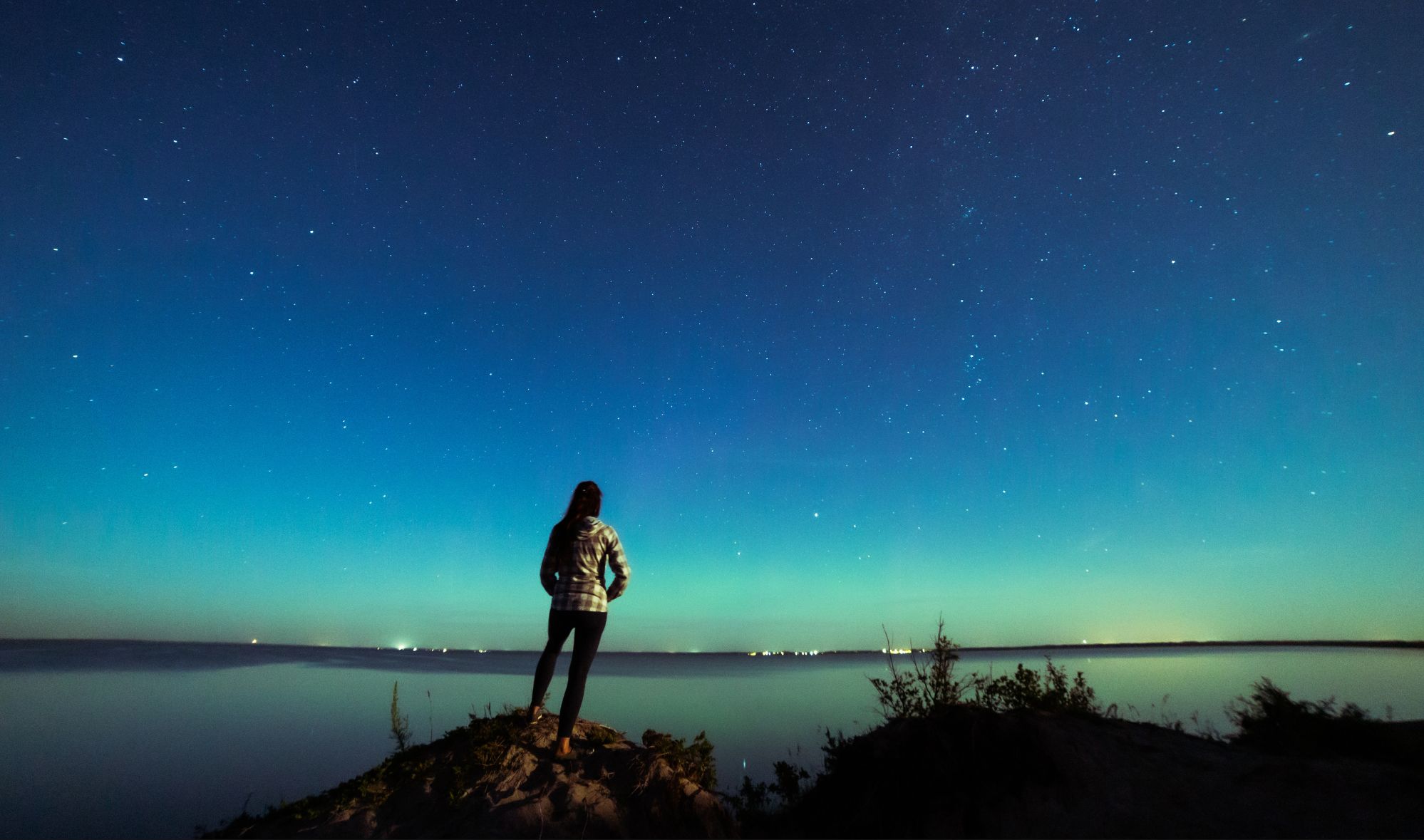 A silhouetted person stands on a rocky bluff overlooking calm water at night, gazing up at a clear sky filled with stars. A soft turquoise glow from the northern lights spreads across the horizon, reflecting faintly on the water below.