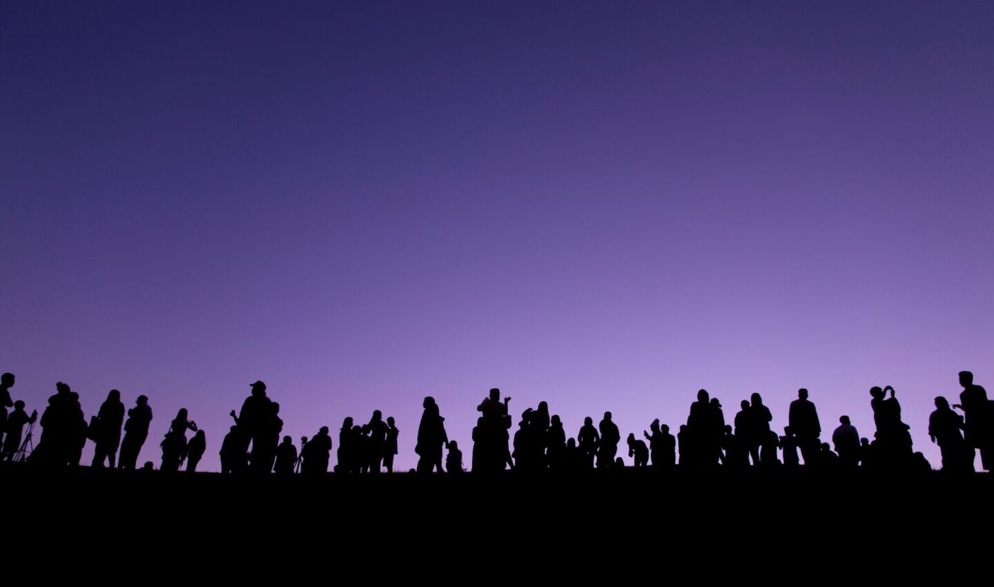 Silhouettes of a large group of people standing along a ridge against a purple twilight sky, some holding cameras and tripods as they watch the horizon.