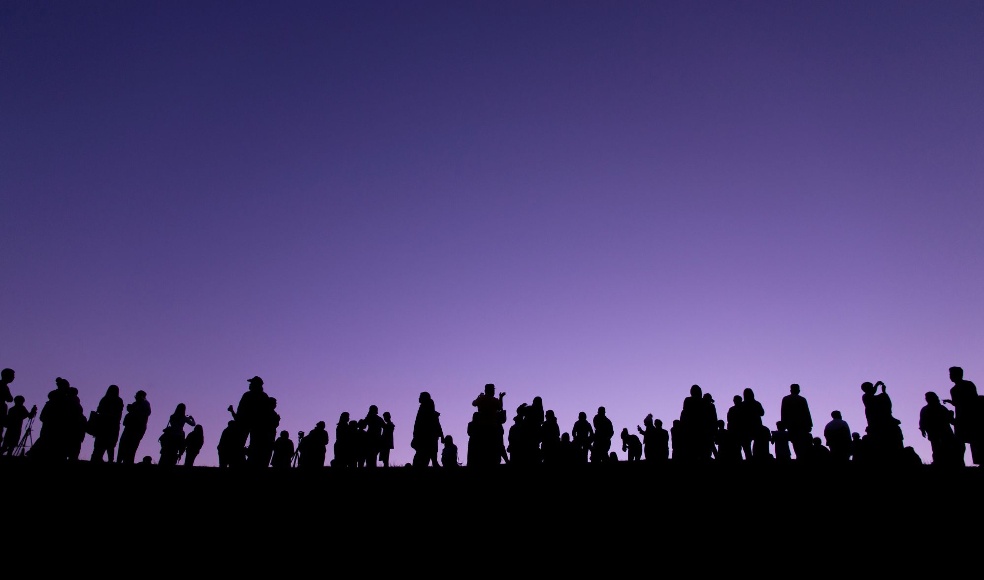 Silhouettes of a large group of people standing along a ridge against a purple twilight sky, some holding cameras and tripods as they watch the horizon.