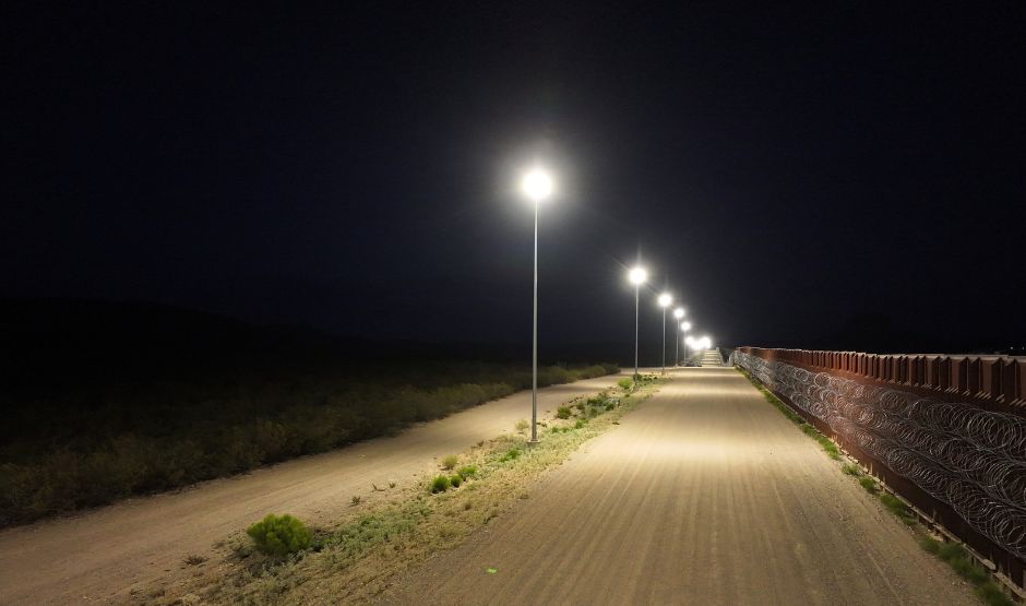 High-powered stadium lights line a dirt road beside the U.S.–Mexico border wall at night, illuminating the desert landscape and cutting through an otherwise dark sky.