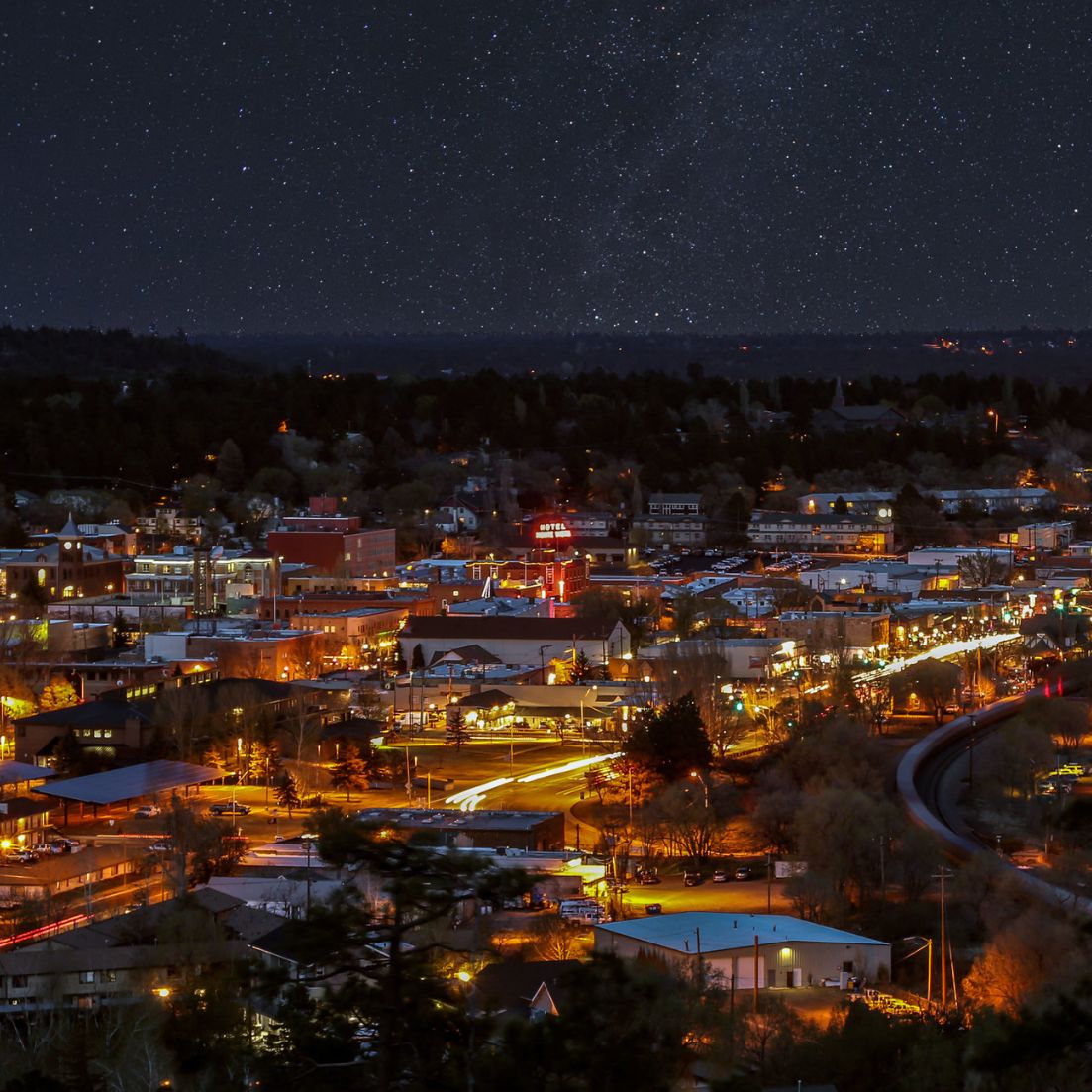 Nighttime view of a small city with warm streetlights and buildings glowing below a star-filled sky, with the Milky Way faintly visible above the horizon.