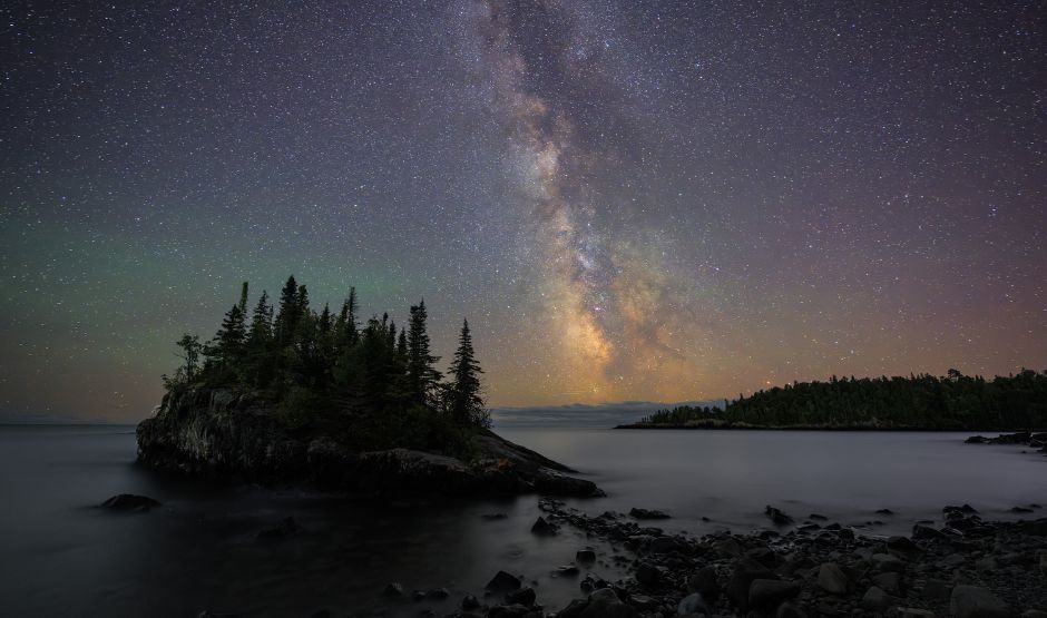 The Milky Way rises over a rocky shoreline and pine-covered island in the Boundary Waters Canoe Area Wilderness, reflected in the calm waters of Duncan Lake under a star-filled night sky.
