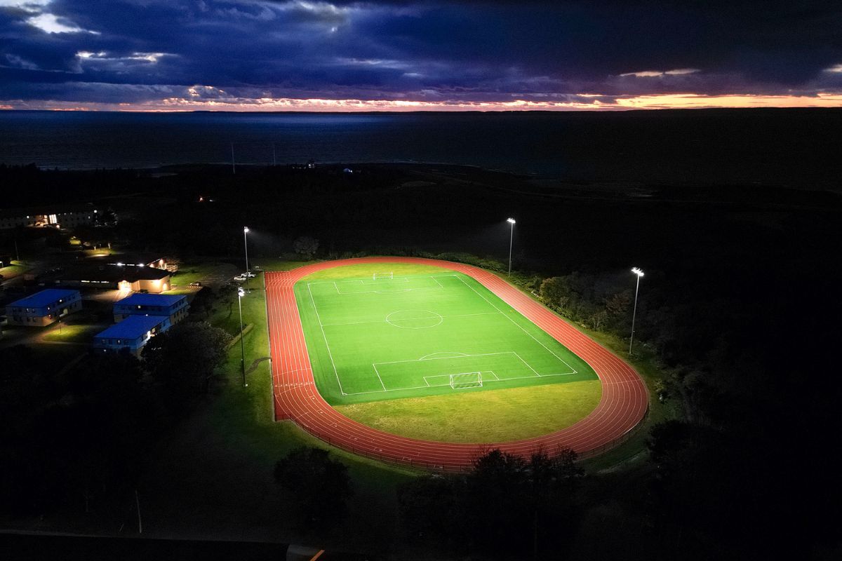 Brightly lit soccer field and running track at night, set against a dramatic coastal sunset and dark storm clouds.