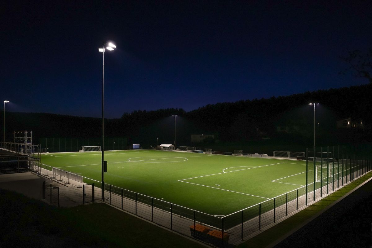 Illuminated soccer field at night, surrounded by fencing and tall floodlights.