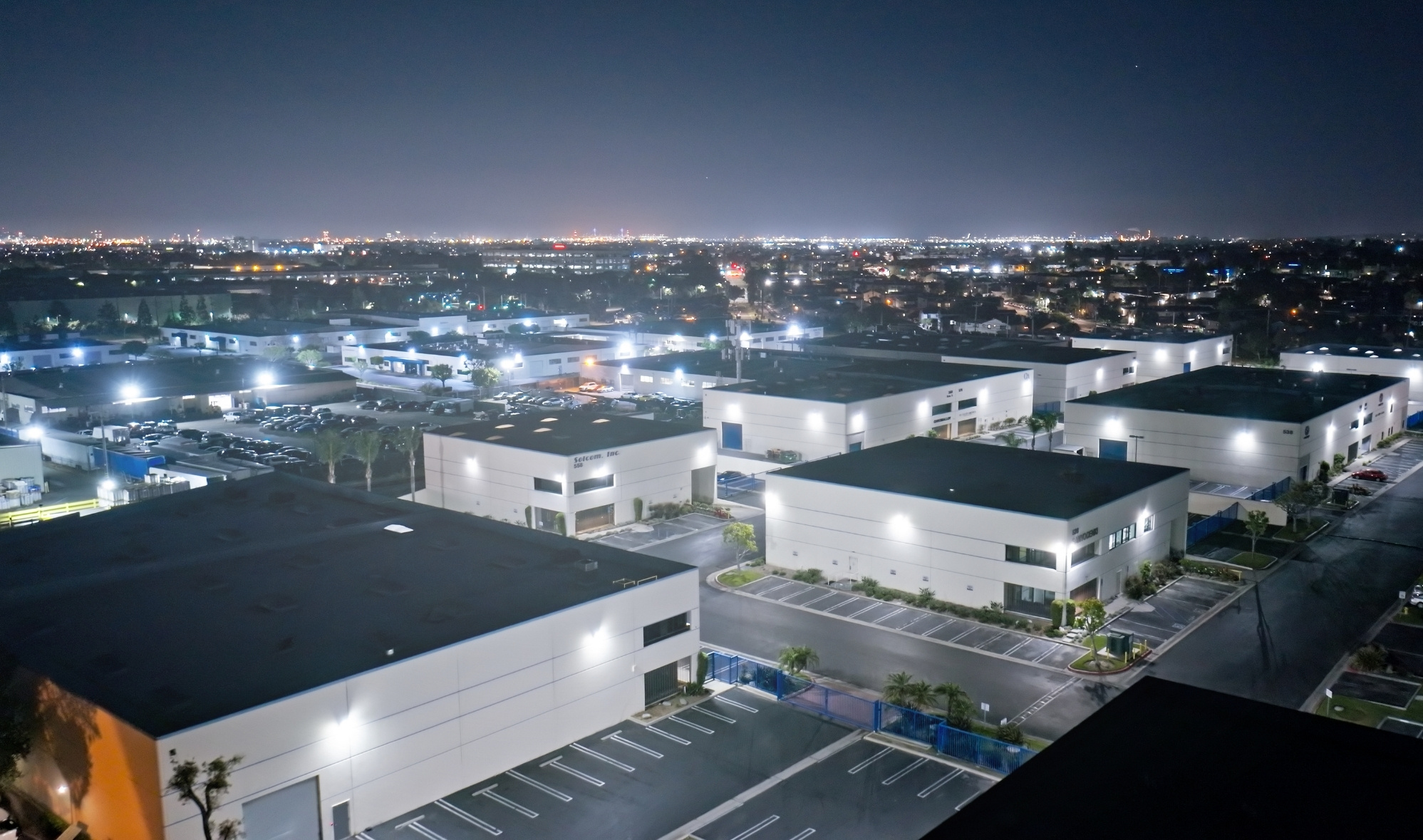 ndustrial park at sunset with bright outdoor lighting creating visible glare above warehouses, roadways, and parking areas.