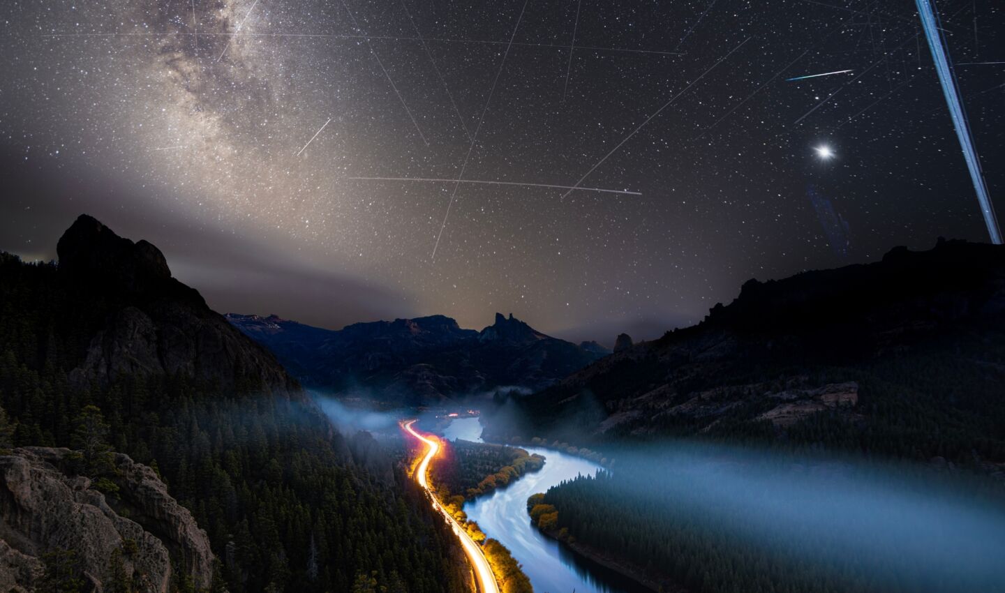 Nighttime view over a rugged mountain valley and winding river beneath the Milky Way. A glowing road traces through mist-covered forests in the foreground, while dozens of bright satellite trails streak across the star-filled sky, intersecting above the landscape. A brilliant planet or star shines near the horizon, highlighting the contrast between the natural night sky and increasing human activity in orbit.