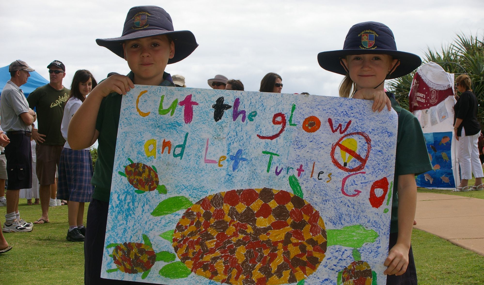 Two schoolchildren wearing wide-brimmed hats hold a colorful handmade poster featuring sea turtles and the message "Cut the glow and let turtles go!" during a community event promoting the protection of sea turtles from light pollution. Several people and educational displays are visible in the background.
