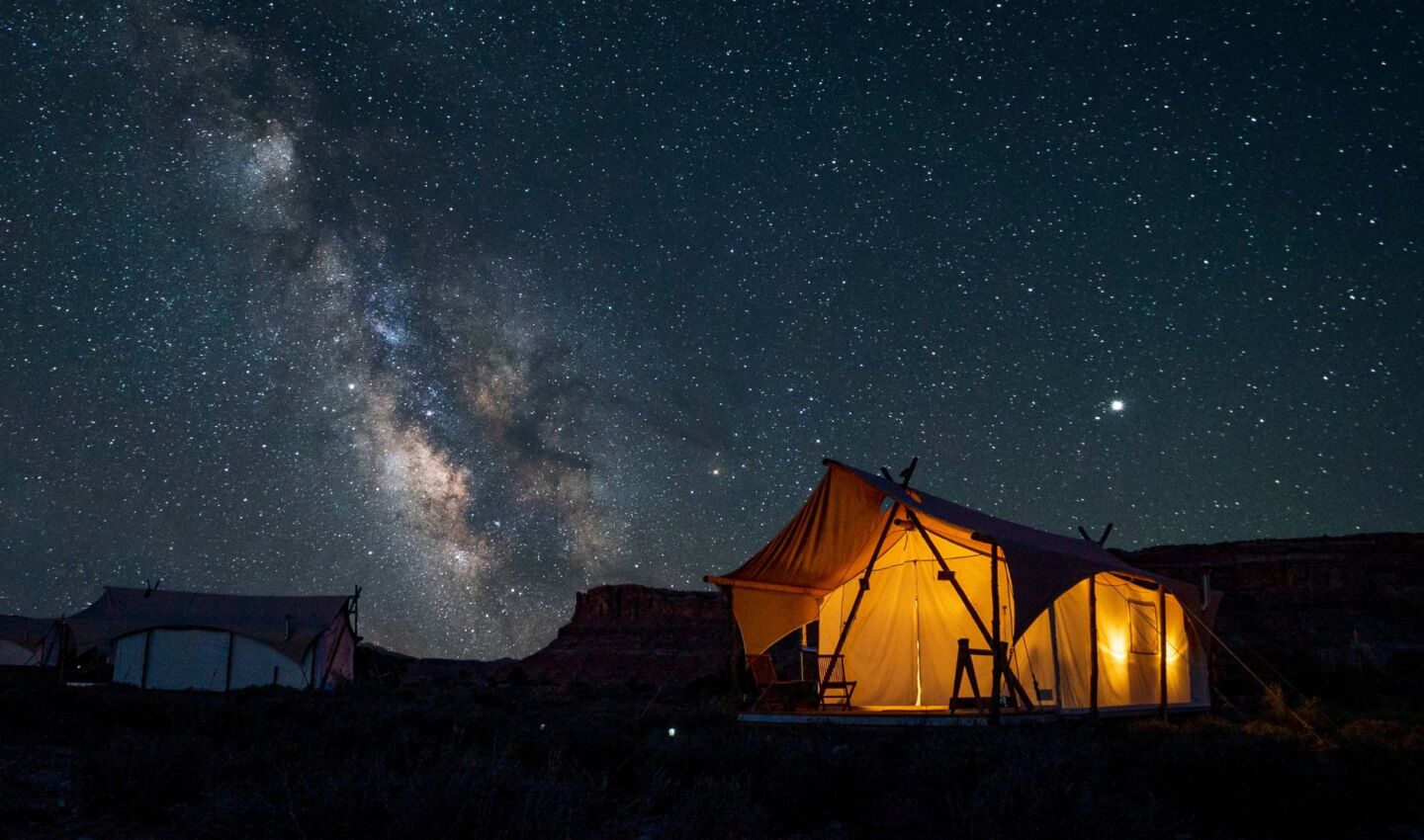 A glowing canvas tent sits beneath a brilliant, star-filled sky in a desert landscape, with the Milky Way rising prominently overhead. Surrounded by rugged mesas and sparse vegetation, the warmly lit tent offers a striking contrast against the dark night, showcasing an immersive stargazing experience in a naturally dark environment.