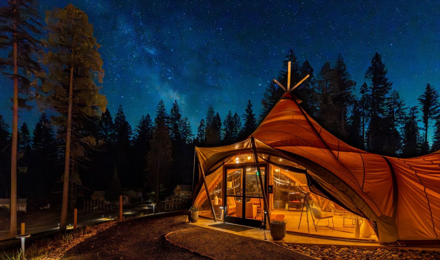 A warmly lit safari-style canvas tent sits beneath a clear, star-filled night sky, with the Milky Way stretching overhead. Surrounded by tall pine trees, the tent's large windows reveal a cozy interior, while soft pathway lighting illuminates the entrance without overpowering the dark nighttime landscape.