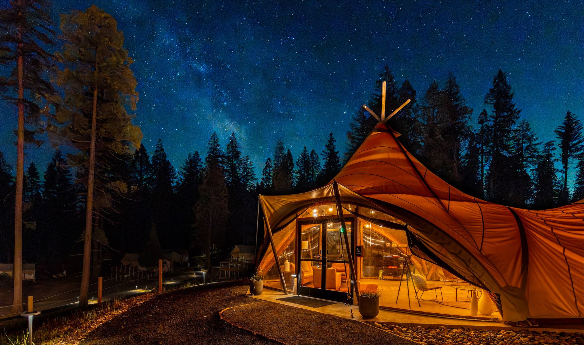 A warmly lit safari-style canvas tent sits beneath a clear, star-filled night sky, with the Milky Way stretching overhead. Surrounded by tall pine trees, the tent's large windows reveal a cozy interior, while soft pathway lighting illuminates the entrance without overpowering the dark nighttime landscape.