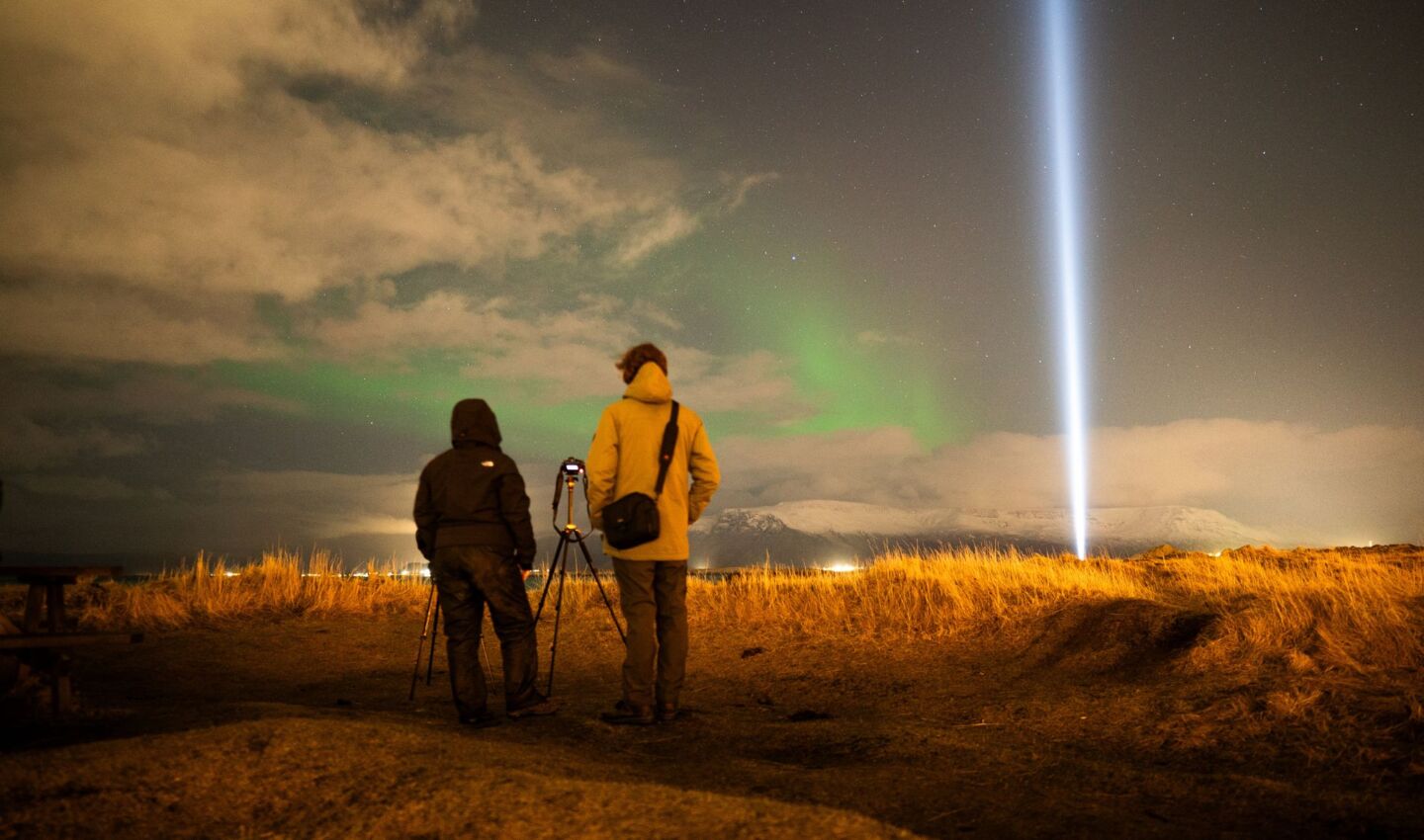 Two people stand beside a camera tripod under a starry sky in Iceland as the faint green glow of the aurora borealis appears above distant mountains. A bright vertical beam of artificial light rises into the sky nearby, illustrating the contrast between natural darkness and light pollution.