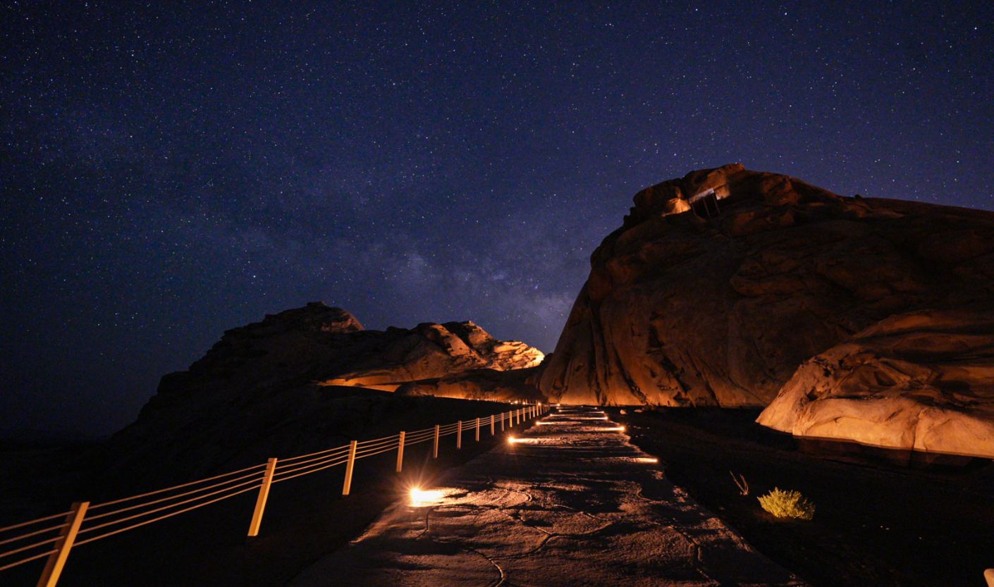 A gently illuminated stone pathway winds through dramatic desert rock formations beneath a star-filled night sky and the Milky Way. Warm, low-level lighting guides the path while preserving the surrounding darkness, creating a tranquil nighttime landscape with minimal light pollution.