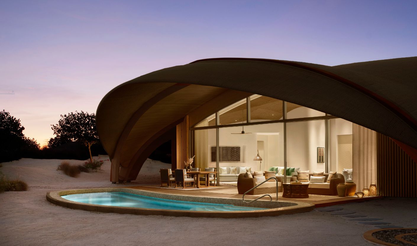 Exterior view of a luxury desert villa at dusk with a sweeping curved roof, floor-to-ceiling glass walls, and a private illuminated pool. Warm interior lighting spills onto the outdoor terrace, surrounded by sand dunes and sparse desert vegetation beneath a soft twilight sky.