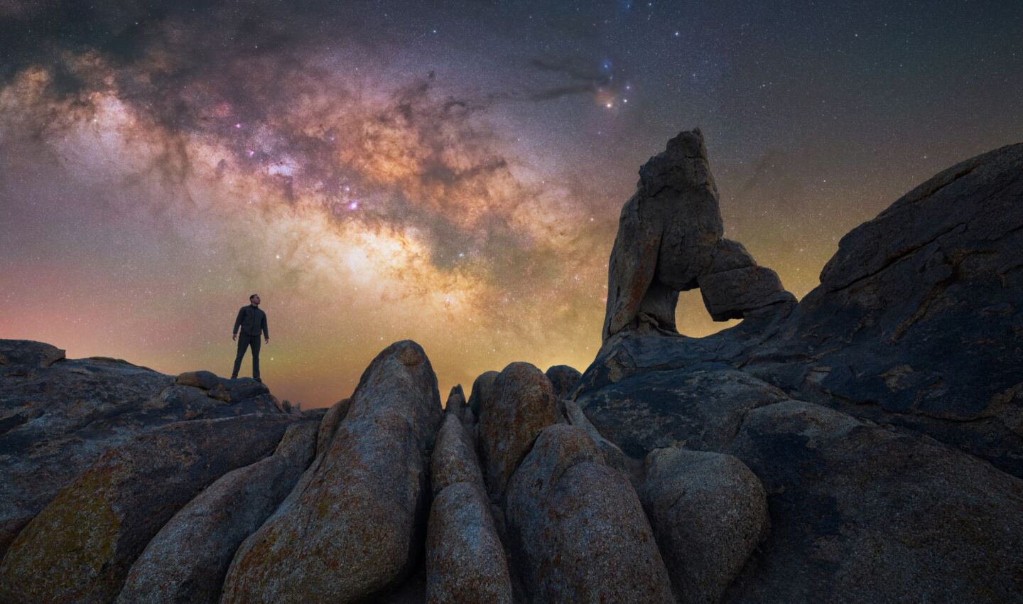 A person stands atop smooth granite boulders beneath the brilliant arch of the Milky Way, gazing toward a dramatic natural rock formation with a window-like opening. The dense star field and colorful galactic core stretch across the night sky, creating a striking contrast with the rugged, dark foreground landscape.