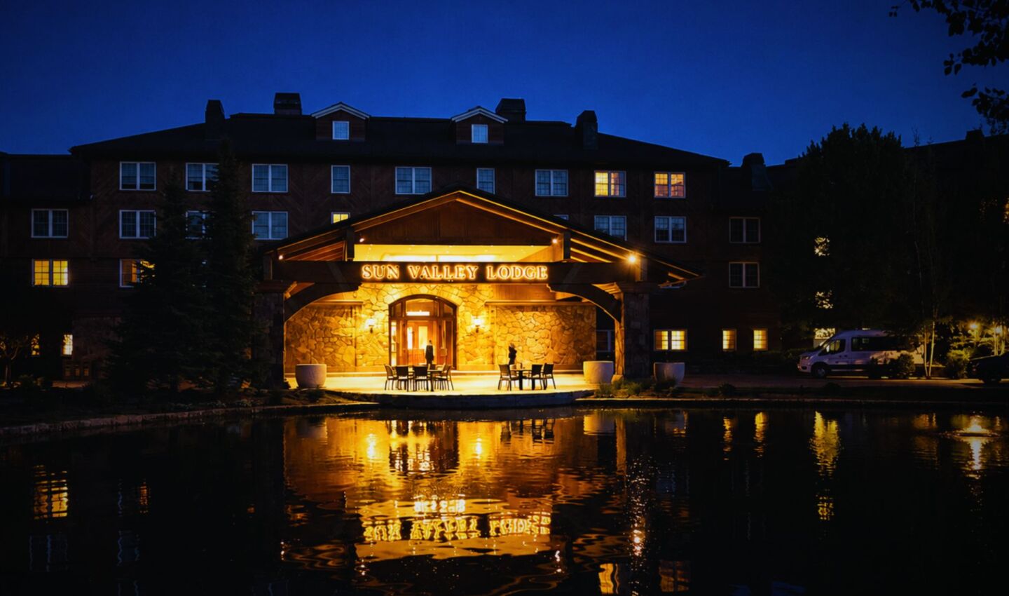 Nighttime view of the entrance to Sun Valley Lodge, warmly illuminated with amber lighting that reflects across a calm pond in the foreground. The multi-story resort rises behind the covered stone entrance beneath a deep blue twilight sky, with only a few guest room windows lit, demonstrating responsible outdoor lighting that preserves the surrounding darkness.