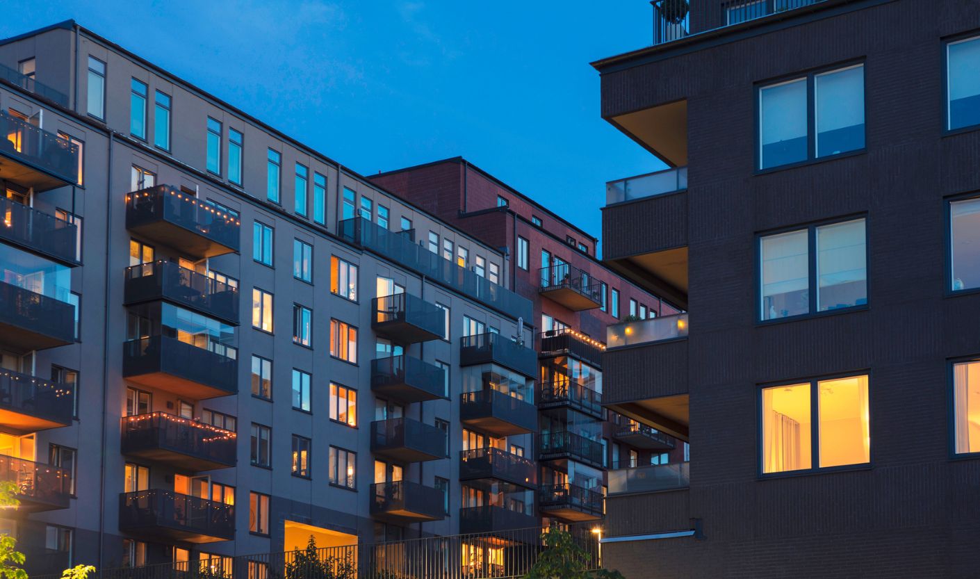 Apartment buildings at dusk with numerous illuminated windows and balconies. Warm indoor lighting contrasts with the blue evening sky, illustrating how residential lighting contributes to urban nighttime light pollution.