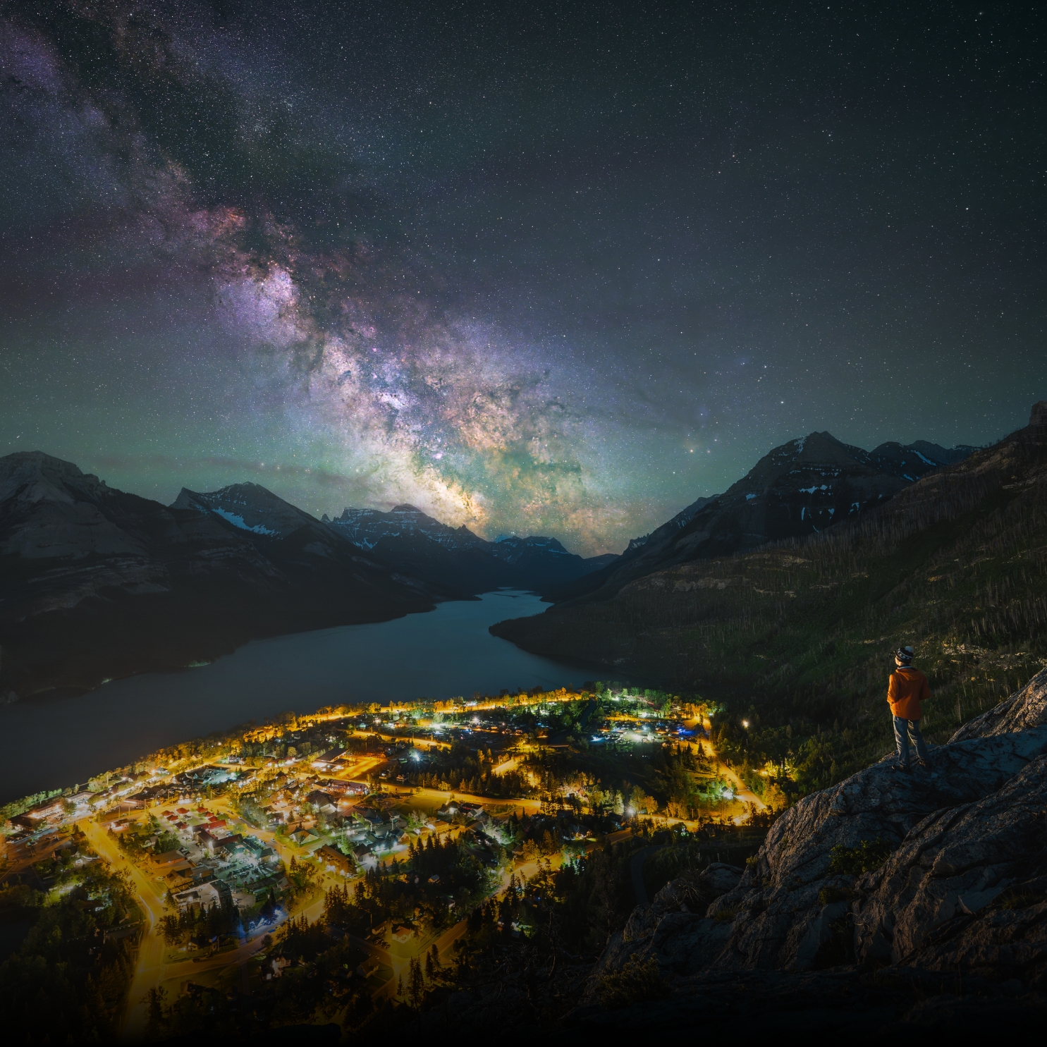 A person in an orange jacket stands on a rocky mountainside overlooking an illuminated lakeside town, with towering mountain peaks and the colorful Milky Way stretching across a star-filled night sky.