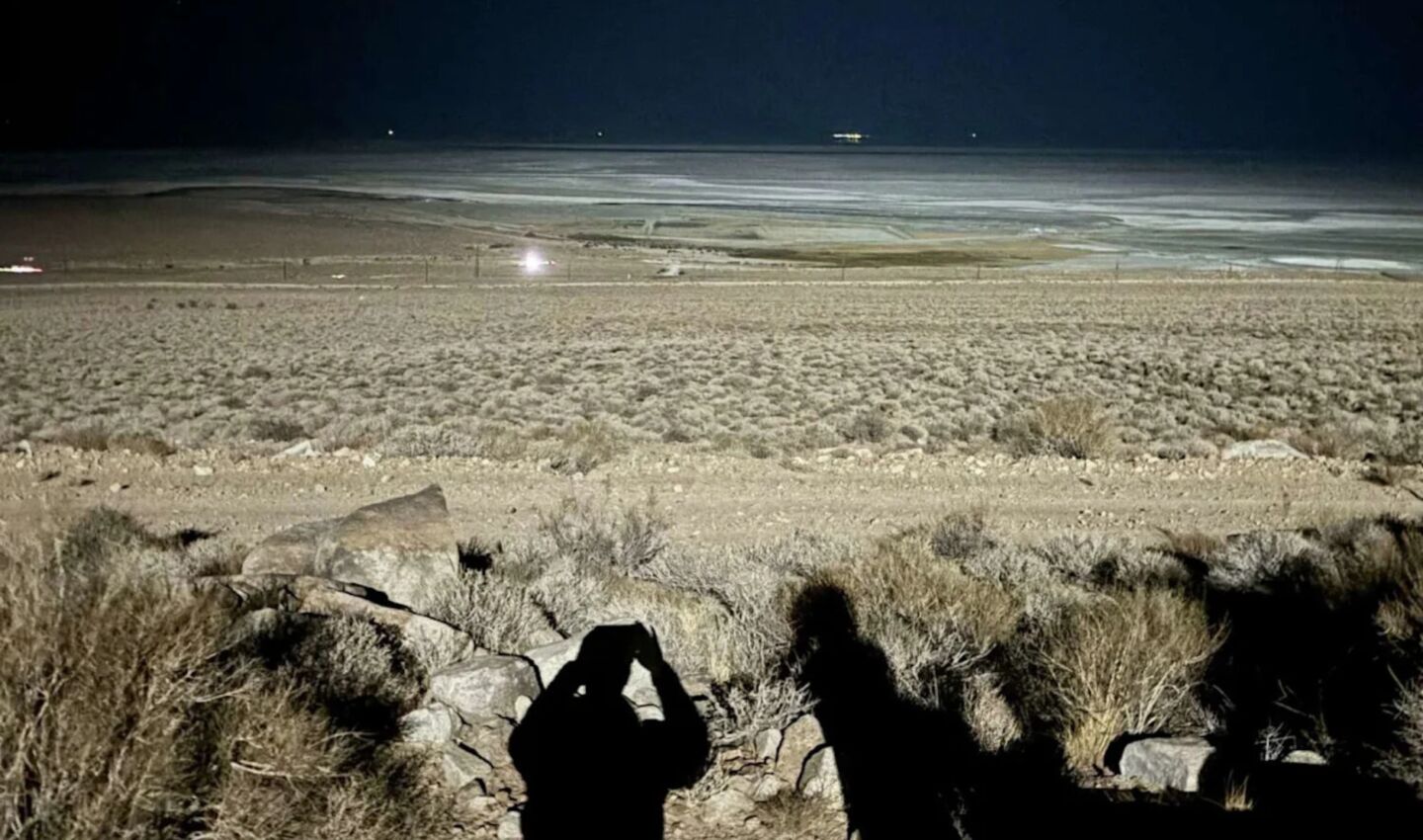 A brightly illuminated desert landscape at night, with sagebrush and rocky terrain in the foreground, distant lights across the valley, and the shadows of two people cast by intense artificial lighting.