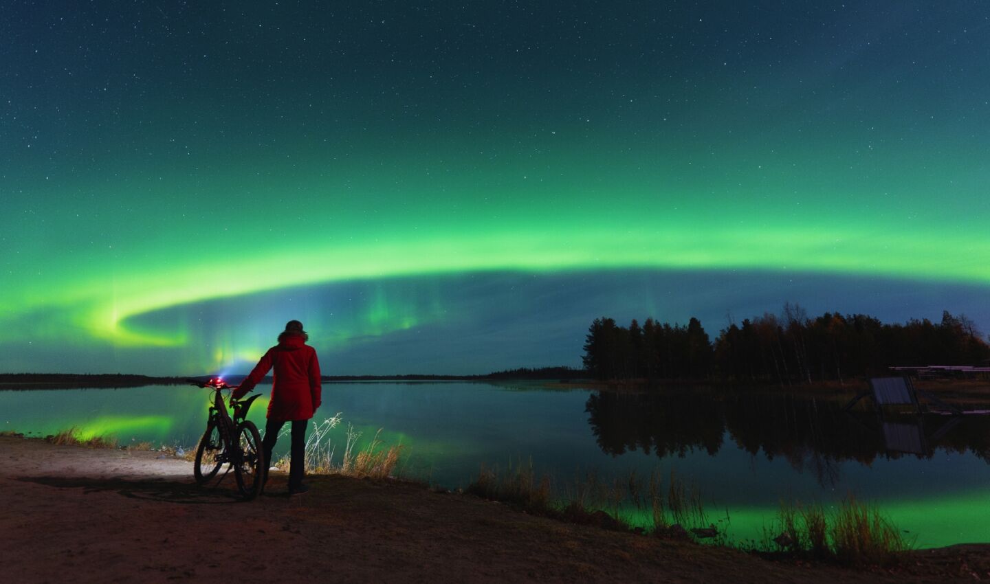 A person in a red jacket stands beside a bicycle on the shore of a calm lake, watching brilliant green northern lights arc across a star-filled night sky and reflect in the water.