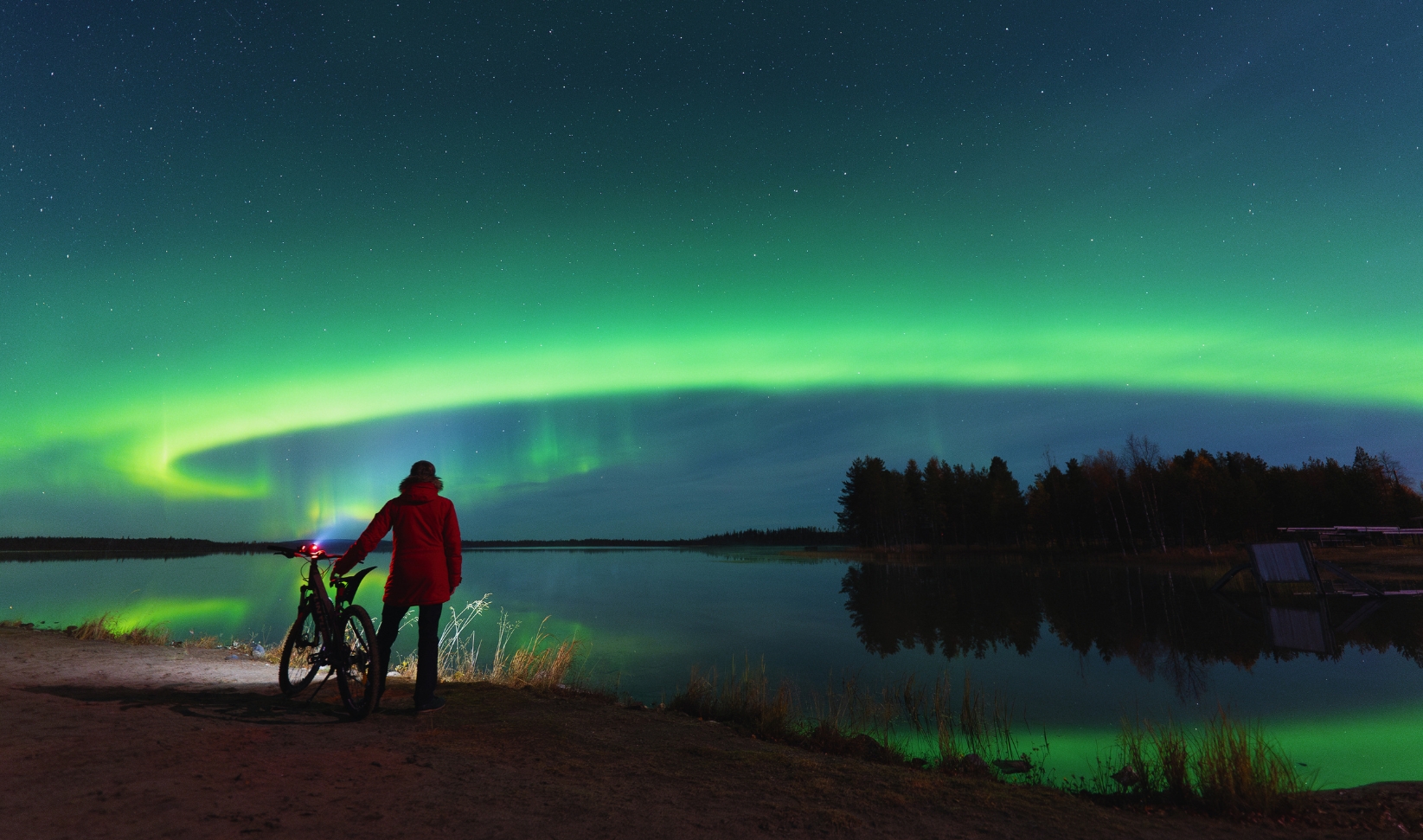 A person in a red jacket stands beside a bicycle on the shore of a calm lake, watching brilliant green northern lights arc across a star-filled night sky and reflect in the water.